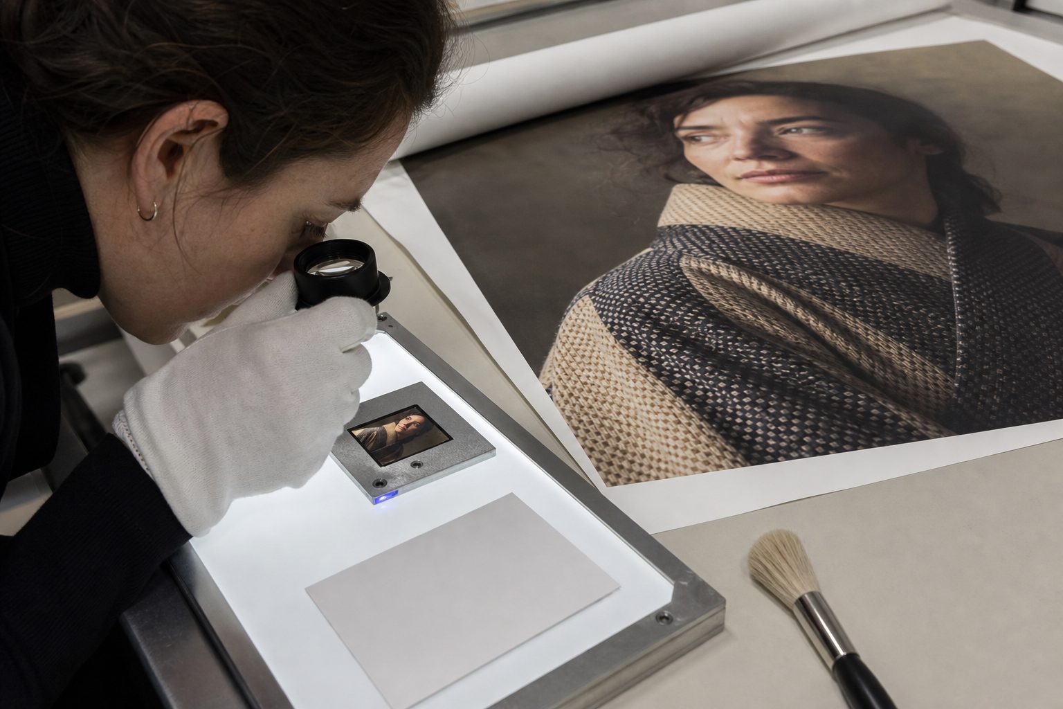 Gloved technician inspects a large portrait print and matching transparency with a loupe on a light table