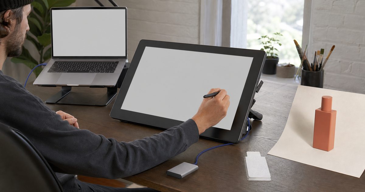Designer works at a pen display beside a laptop, product bottle print, cables, and an external drive
